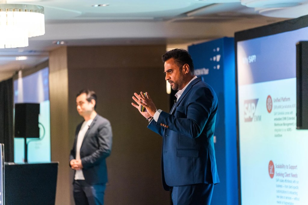 A business presentation at the SAP NOW AI Tour in Singapore, featuring a speaker gesturing while discussing digital transformation, with another person in the background and a presentation screen displaying SAP-related information.